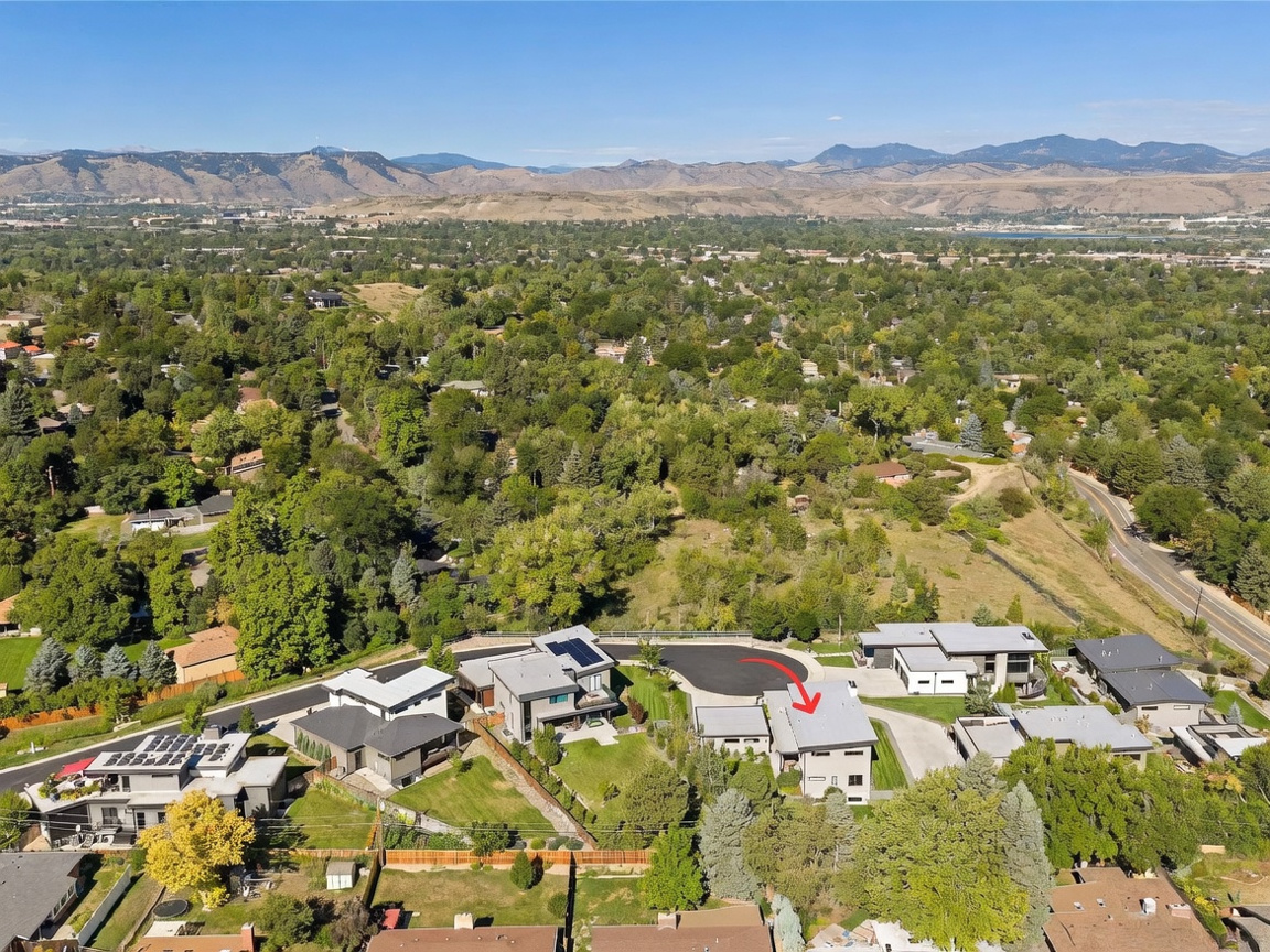 This high-angle aerial view captures a modern residential neighborhood nestled against a backdrop of rolling hills and mountains. The scene highlights several contemporary homes with flat or low-pitched roofs, manicured lawns, and paved driveways, with a red arrow pointing toward a specific property. The surrounding landscape is lush with mature trees, creating a serene and suburban atmosphere.