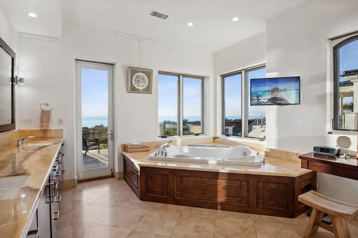 This luxurious primary bathroom features a large soaking tub set into a stone-topped platform, positioned to take advantage of expansive ocean views through large windows. A long vanity with dual sinks and stone countertops runs along the left wall, complemented by neutral-toned tile flooring. The space is bright and airy, with a glass door providing direct access to an outdoor patio area.