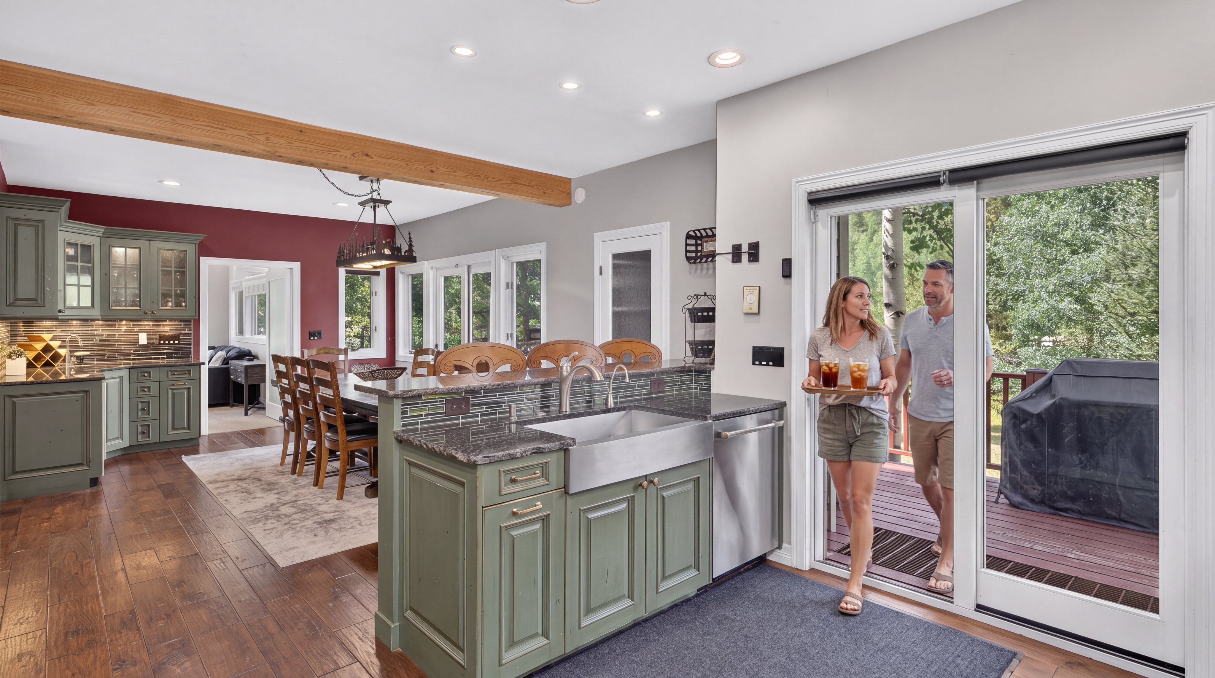 This interior view showcases an open-concept kitchen and dining area featuring Sage green cabinetry, dark granite countertops, and a farmhouse-style stainless steel sink. A large wooden dining table sits adjacent to the kitchen, while a glass sliding door connects the space to an outdoor deck where a couple is entering with beverages. The home's warm aesthetic is complemented by hardwood flooring, a striking red accent wall, and exposed wooden ceiling beams.