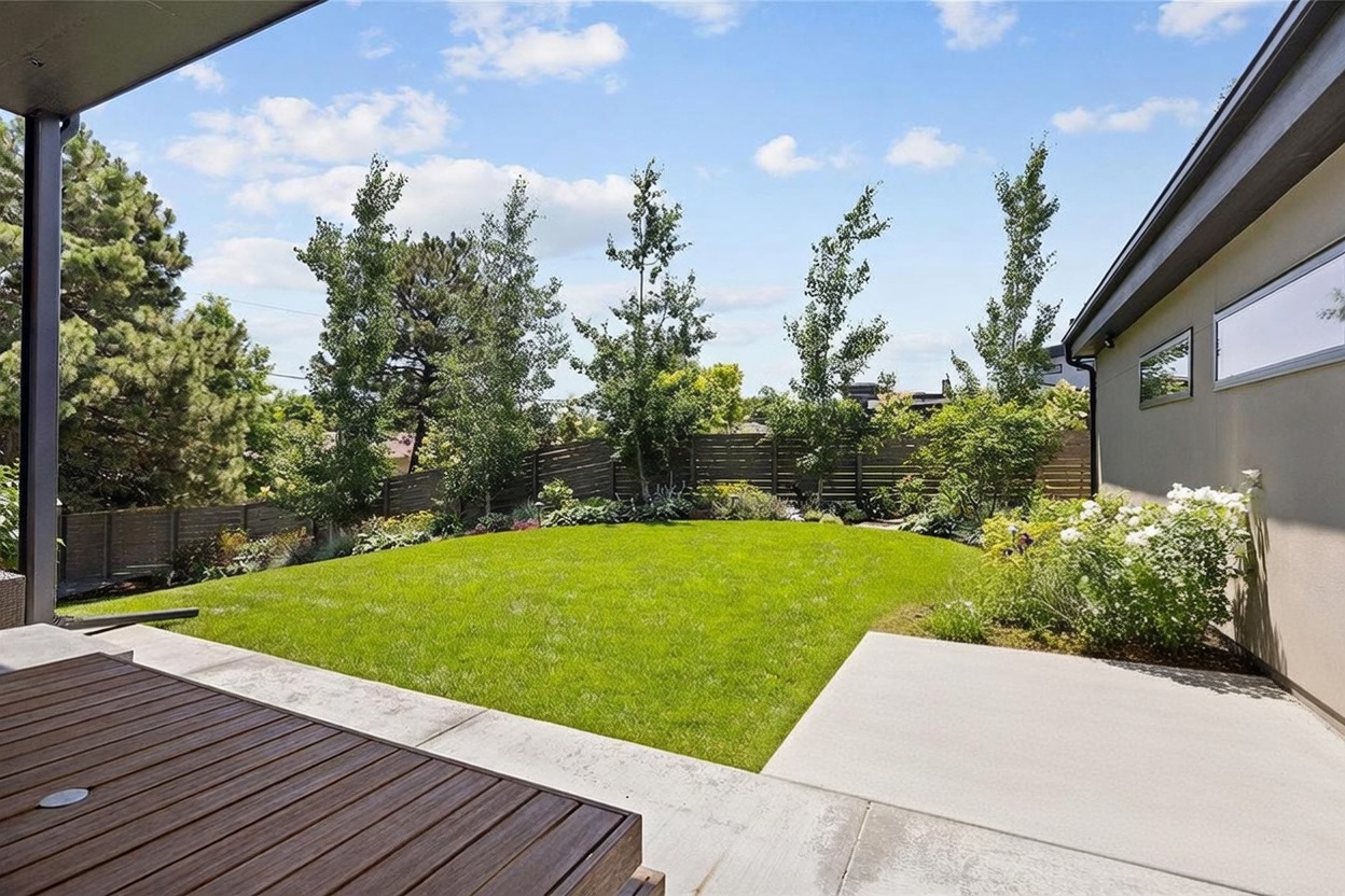 This image captures a well-manicured backyard viewed from a covered patio area. The space features a lush green lawn bordered by vibrant flower beds, mature trees, and a horizontal wood privacy fence. The perspective highlights the seamless transition between the indoor-outdoor living space and the private, landscaped garden.