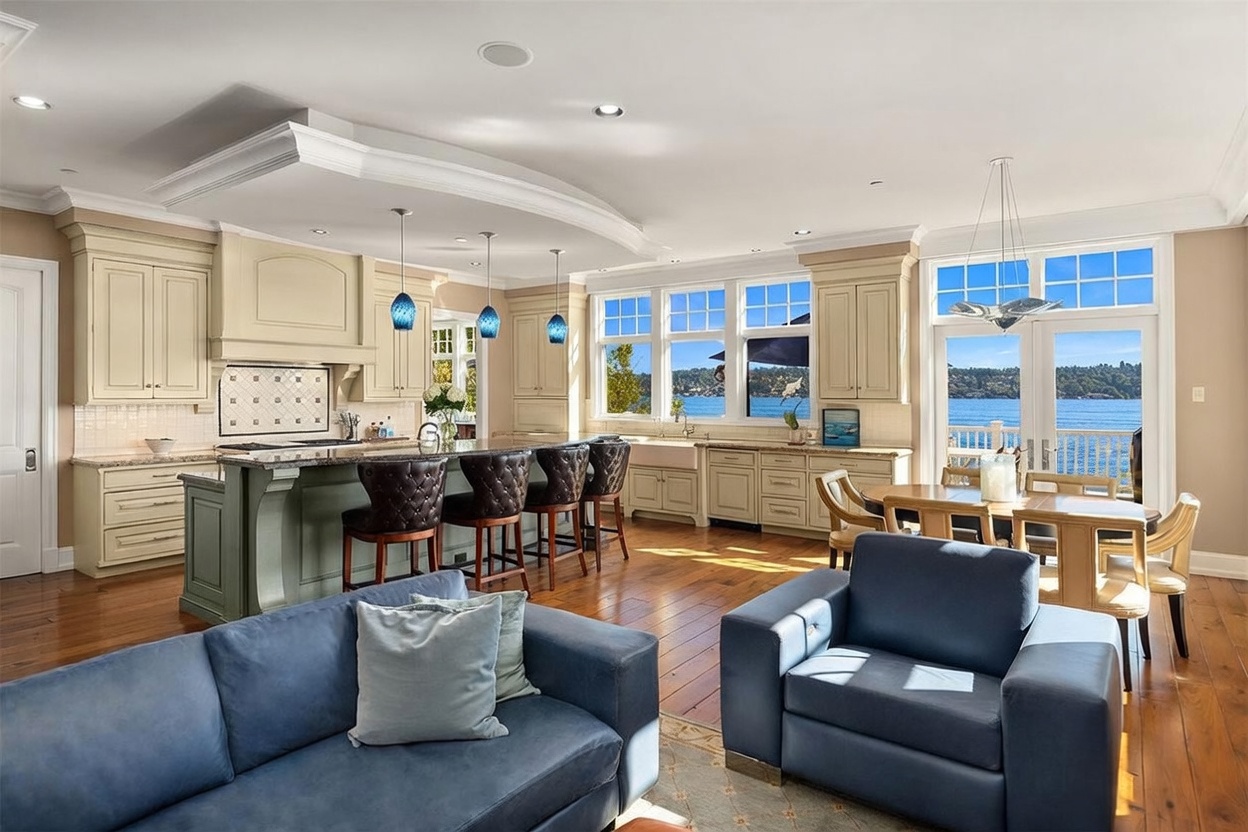 This spacious, open-concept kitchen features a large sage-green island with dark leather bar stools, complemented by cream-colored cabinetry and warm hardwood flooring. The room is bathed in natural light from expansive windows that offer a stunning, unobstructed view of the water, creating a seamless connection between the interior and the outdoors. A dining area is conveniently situated nearby, making this an ideal space for both casual meals and elegant entertaining.