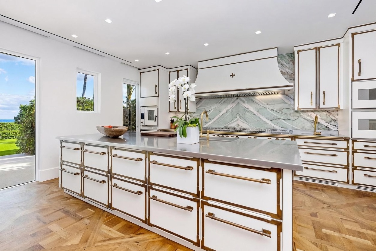 This sophisticated kitchen features a large central island with a metallic countertop and extensive white cabinetry accented by brass-toned hardware. A striking green and white veined stone backsplash serves as a focal point behind the range, complemented by a custom-designed range hood. The room is finished with light-toned herringbone wood flooring and opens through a glass door to a view of the outdoors.