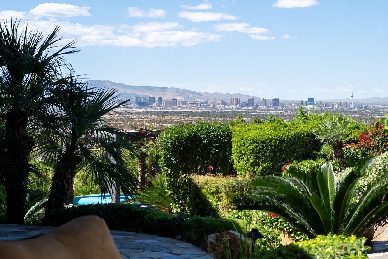 This elevated patio offers a breathtaking, panoramic view of the Las Vegas skyline set against a backdrop of distant mountains. The foreground features lush, manicured landscaping with vibrant green hedges and palm trees, framing a glimpse of a private pool area below. The stone-paved terrace provides a serene vantage point, capturing the essence of luxury desert living with a seamless blend of natural beauty and urban scenery.