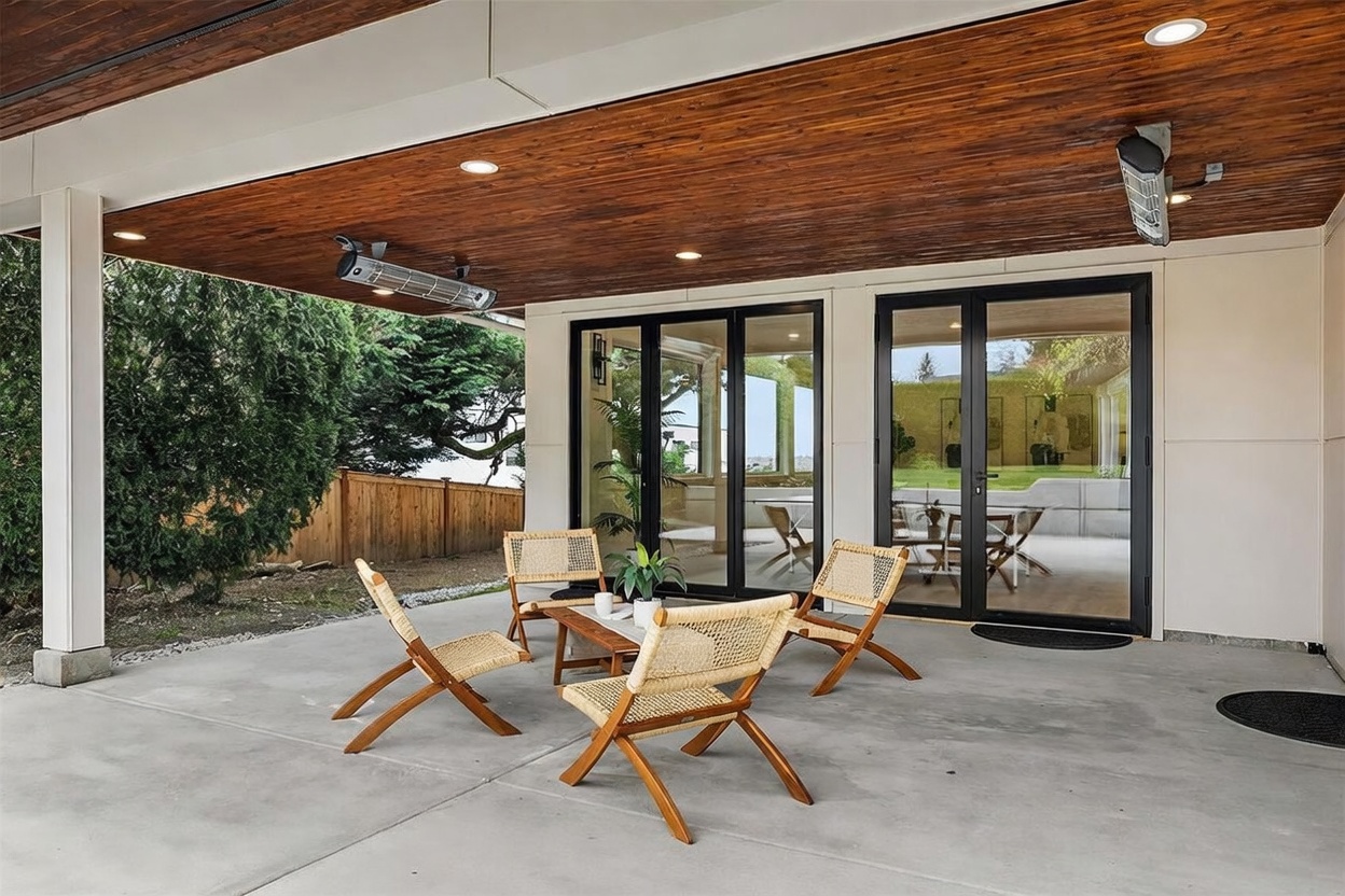 This covered patio features a concrete floor and a warm, wood-paneled ceiling with recessed lighting and mounted outdoor heaters. The space is furnished with four light-wood chairs with woven seats arranged around a small coffee table, providing a comfortable outdoor seating area. Large glass doors with black frames lead into the home, creating a seamless transition between the indoor and outdoor living spaces.