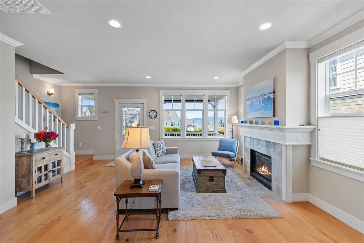This inviting living room features warm light wood flooring and a neutral color palette, centered around a cozy fireplace with a decorative tile surround and a white mantel. A comfortable sofa and two accent chairs are arranged on a textured area rug, while large windows provide ample natural light and a view of the outdoors. To the left, a wooden staircase with white railings leads to an upper level, and a rustic console table adds a touch of character to the space.