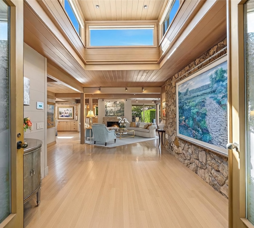 This grand entryway features a dramatic vaulted ceiling with a central skylight that floods the space with natural light. The foyer transitions from a large, textured stone-tiled floor into a spacious living area defined by light wood flooring and a prominent stone-clad wall adorned with a large landscape painting. The open-concept layout creates a seamless flow, highlighting the architectural volume and the sophisticated, airy atmosphere of the home.