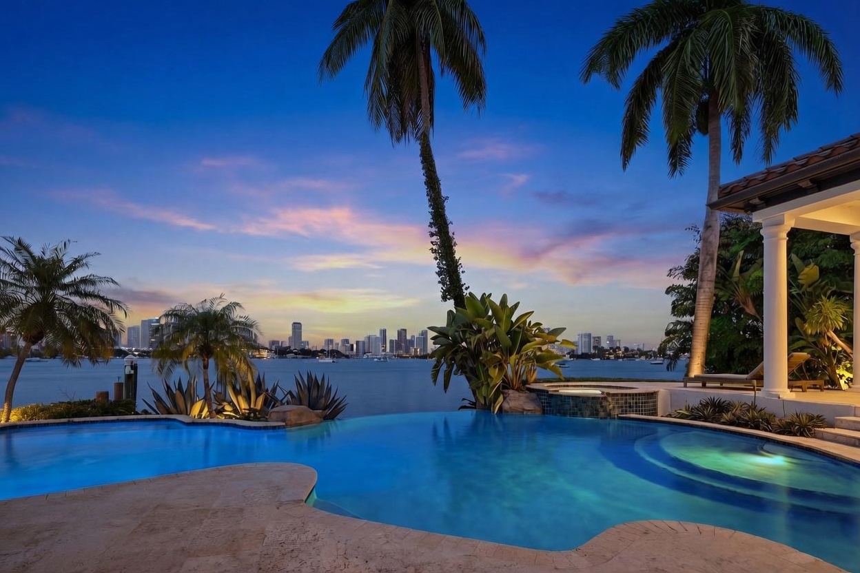 This stunning waterfront property features a custom-shaped infinity-edge pool that seamlessly blends with the bay, offering a panoramic view of the city skyline at sunset. The poolside area is finished with light-toned stone decking and includes a raised spa, lush tropical landscaping with tall palm trees, and a covered patio space with classic columns. The scene captures a luxurious, serene atmosphere perfect for evening relaxation and entertaining.