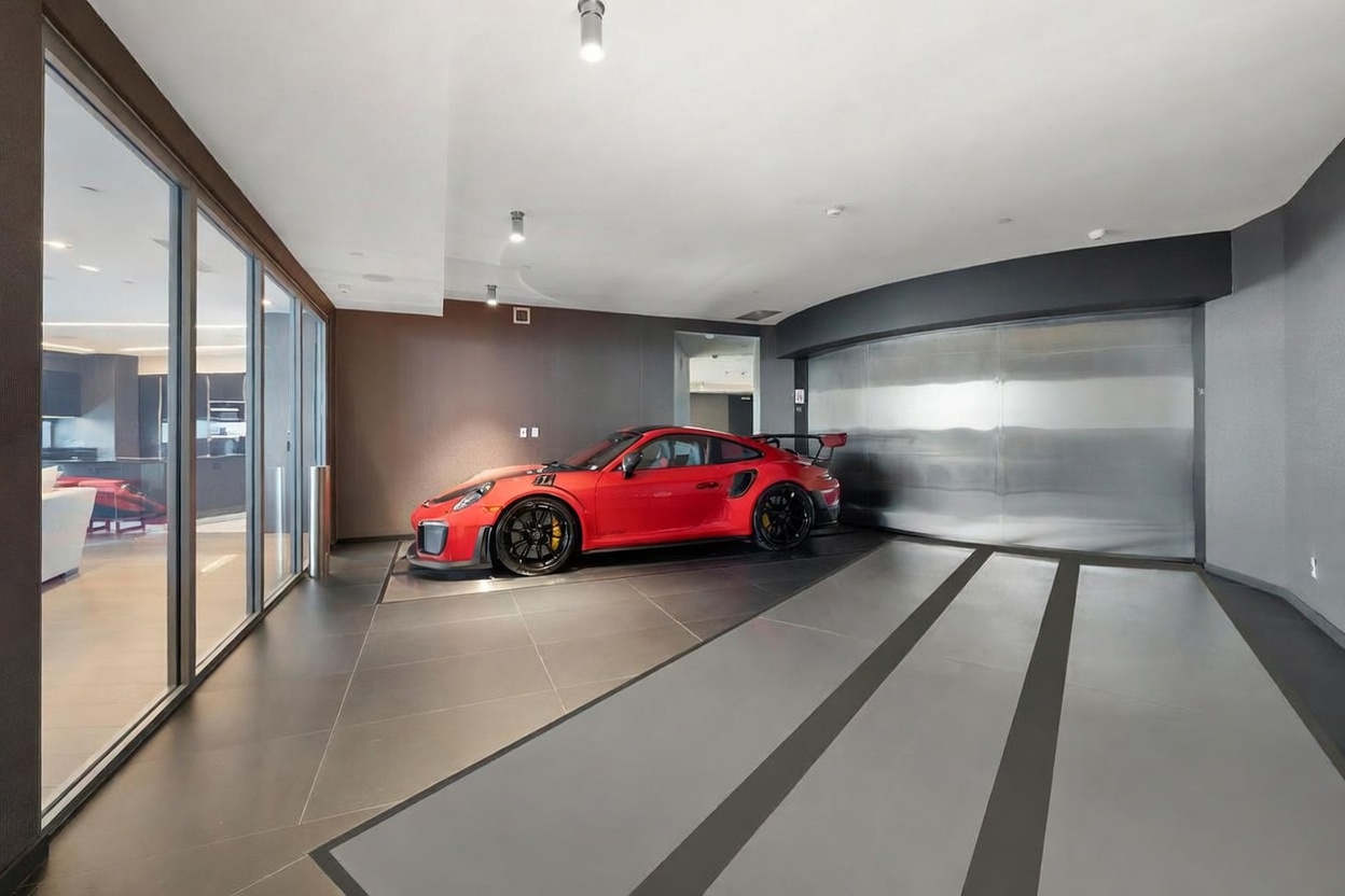 This high-end, modern private garage features a sleek, minimalist design with dark tile flooring and a large, metallic industrial-style door. A vibrant red sports car is parked on a dedicated display platform, serving as the focal point of the space. To the left, a glass wall provides a view into an adjacent interior room, enhancing the sophisticated and gallery-like atmosphere of the garage.