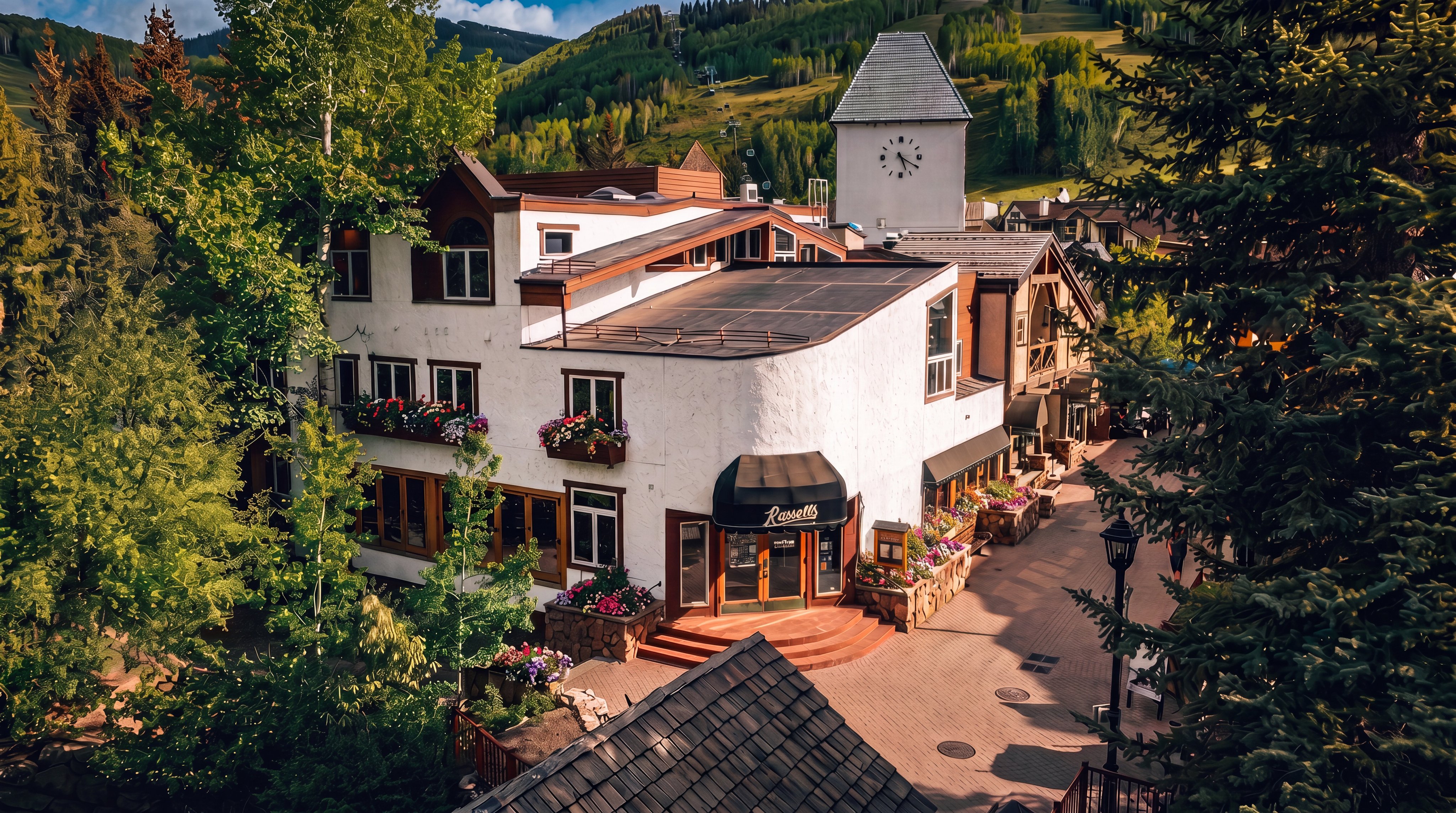 This elevated perspective captures a charming multi-story building in Vail, Colorado, featuring a distinctive clock tower and a prominent storefront for 'Russell's'. The architecture blends rustic mountain elements with stucco walls and dark wood trim, set against a backdrop of lush green ski slopes. The scene includes a pedestrian walkway, mature pine trees, and a clear view of the surrounding alpine landscape.
