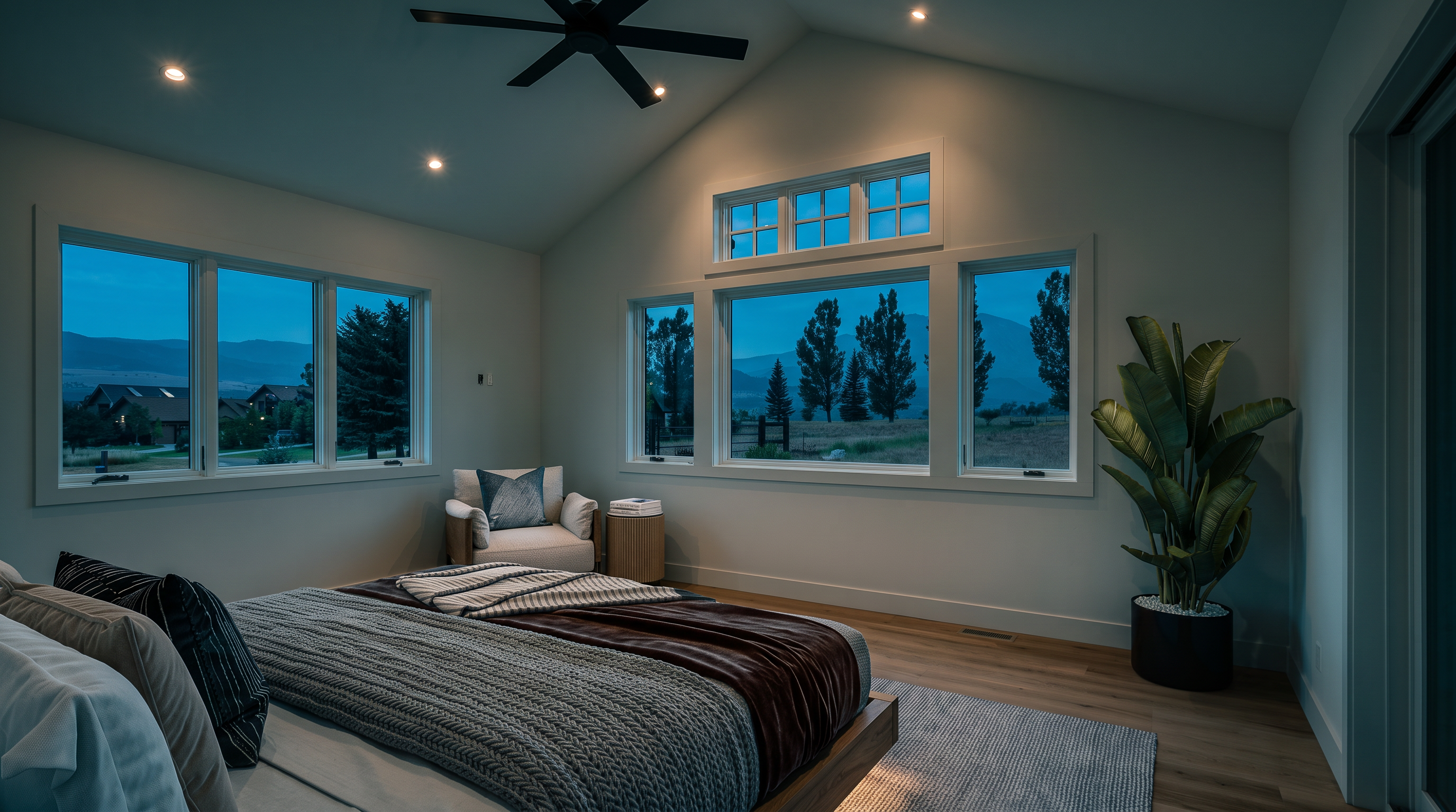 This bright and spacious primary bedroom features a modern, airy design with vaulted ceilings and expansive windows that frame picturesque views of the surrounding landscape. The room is decorated in a neutral, calming color palette, complemented by light hardwood flooring and a stylish, minimalist aesthetic. A large indoor plant and cozy seating area near the windows add warmth and tranquility to this inviting space.