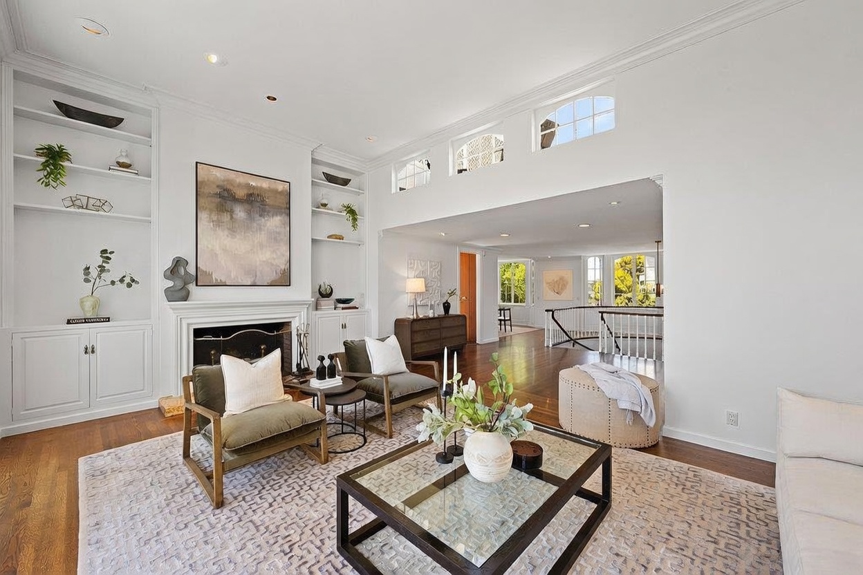 This bright and airy living room features a central fireplace with a classic mantel, flanked by built-in shelving units. The space is furnished with two olive-toned armchairs, a glass-topped coffee table, and a neutral-colored ottoman, all set upon a textured area rug. The room opens into a hallway area with hardwood floors, creating an inviting and open-concept feel.