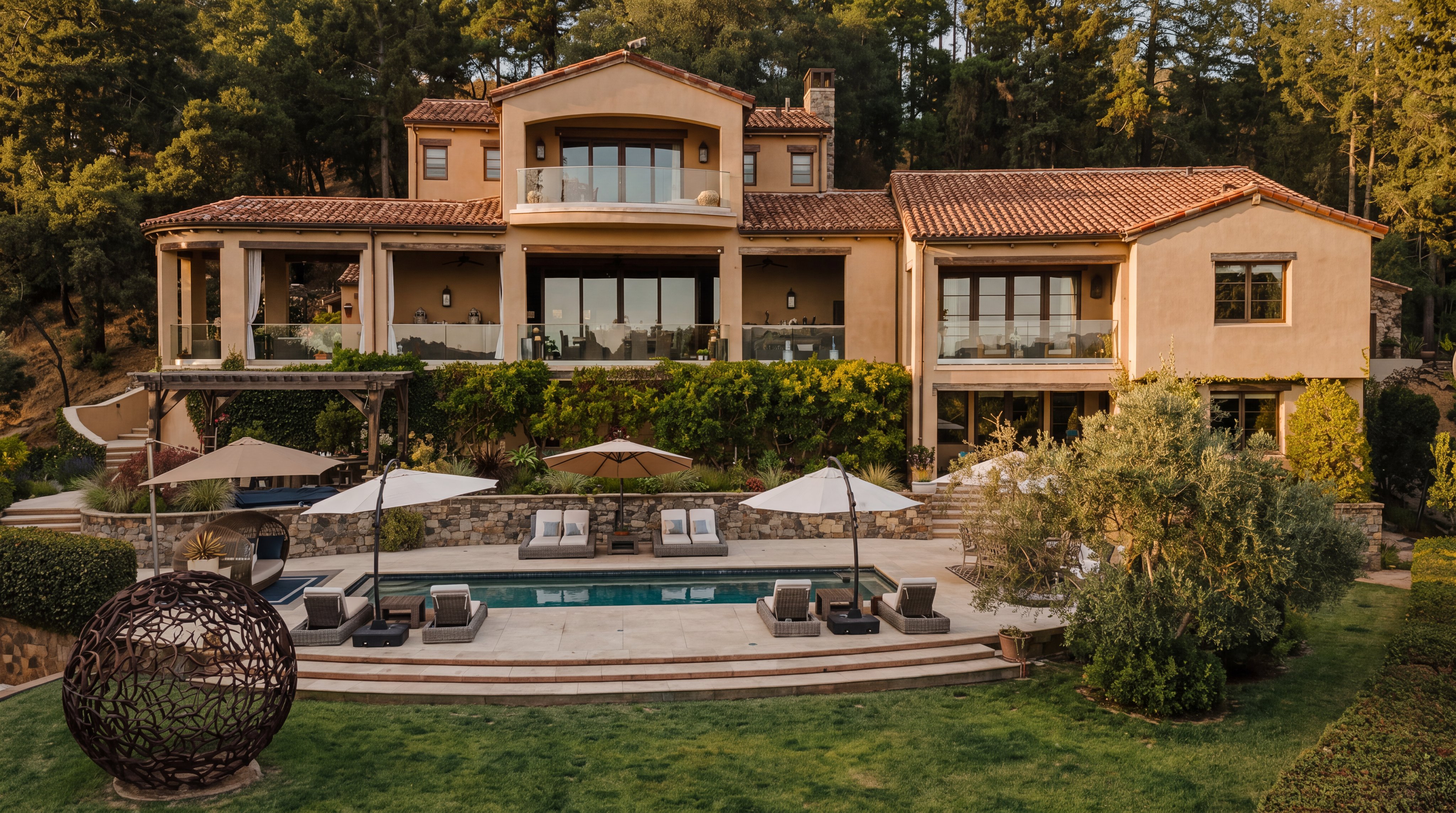 This expansive Mediterranean-style estate features a multi-level rear facade with warm-toned stucco walls and a classic terracotta tile roof. The property boasts a large swimming pool surrounded by a stone patio, multiple outdoor seating areas with umbrellas, and a lush, manicured lawn. The home is nestled against a backdrop of dense, mature trees, creating a private and serene atmosphere.