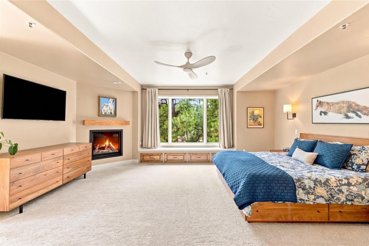 This spacious primary bedroom features a neutral color palette, plush carpeting, and a large window with a built-in bench seat overlooking trees. The room is anchored by a wooden bed frame with blue patterned bedding and a matching dresser, while a fireplace with a wooden mantel adds warmth to the space. The perspective is a wide-angle shot from the doorway, capturing the entire layout including the ceiling fan and wall art.