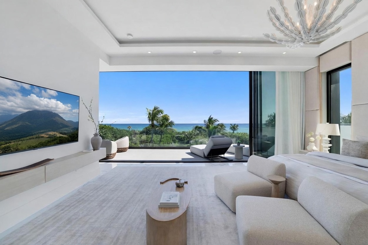 This luxurious primary bedroom features a minimalist, modern aesthetic with a neutral color palette and expansive floor-to-ceiling glass walls that open onto a private balcony overlooking the ocean. The room is furnished with a plush bed, two contemporary lounge chairs, and a sleek, low-profile media console beneath a large wall-mounted television. A unique, sculptural chandelier hangs from the ceiling, adding a sophisticated focal point to the serene, light-filled space.