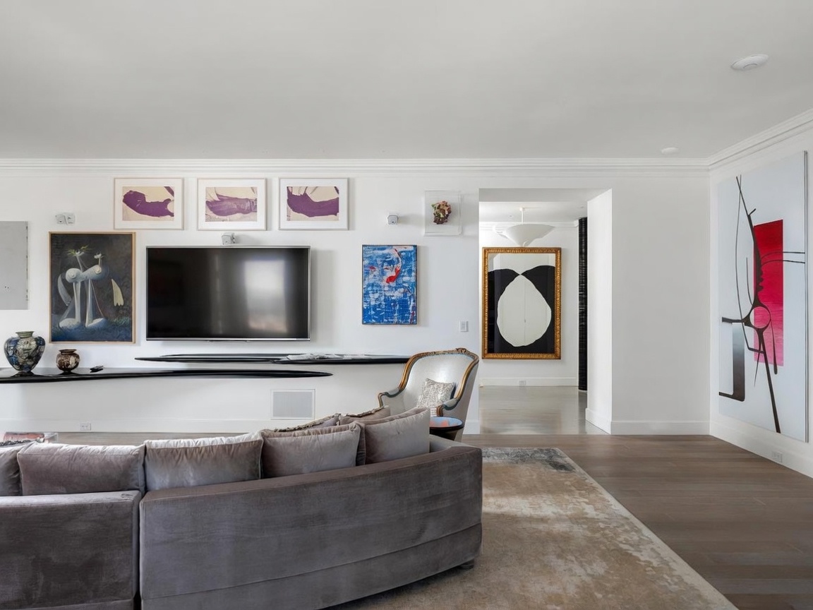 This sophisticated living room features a large, curved grey sectional sofa positioned on a textured area rug, facing a wall adorned with a gallery of modern art and a mounted television. A sleek, minimalist black shelf extends horizontally across the wall, providing a contemporary display surface beneath the screen. The space flows into an adjacent hallway, where a striking black and white abstract painting serves as a focal point, all set against crisp white walls and warm wood flooring.