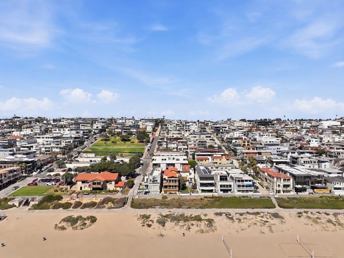This high-angle aerial view captures a coastal neighborhood situated directly along a sandy beach. The scene features a dense collection of multi-story residential buildings, a public park with green space, and a wide expanse of beach with volleyball nets in the foreground. The perspective provides a comprehensive look at the property's prime location and proximity to the ocean.
