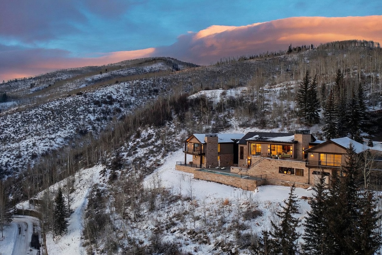 This stunning aerial perspective captures a luxurious mountain estate nestled into a snowy, forested hillside at dusk. The multi-level residence features a sophisticated blend of stone and wood siding, with expansive glass walls that glow warmly against the twilight sky. A sleek infinity-edge pool extends from the stone terrace, overlooking the vast, rugged landscape of the surrounding valley.