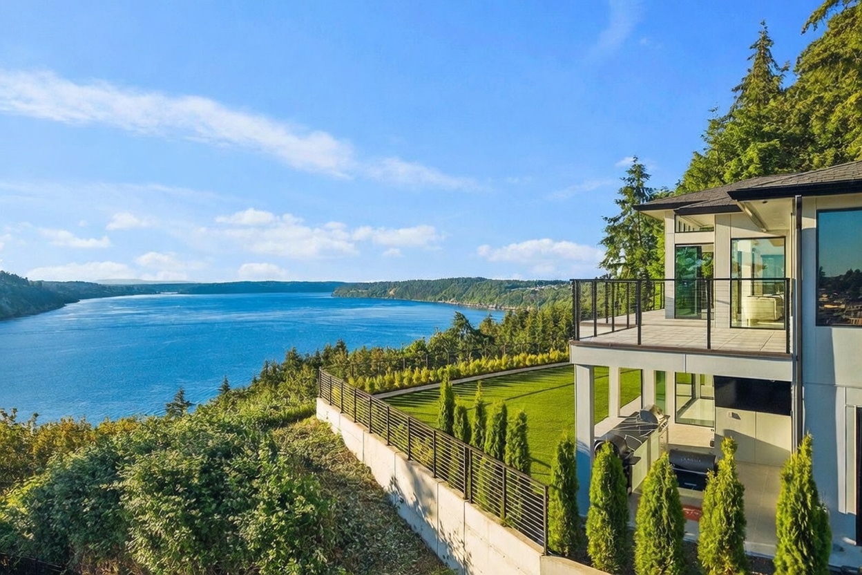 This elevated perspective captures the rear of a modern home overlooking a vast, serene body of water. The property features a spacious upper-level deck with glass railings and a lower-level covered patio area equipped with a grill, all set against a backdrop of lush green trees and a manicured lawn. The architectural design emphasizes clean lines and expansive glass, creating a seamless connection between the indoor living spaces and the breathtaking natural scenery.