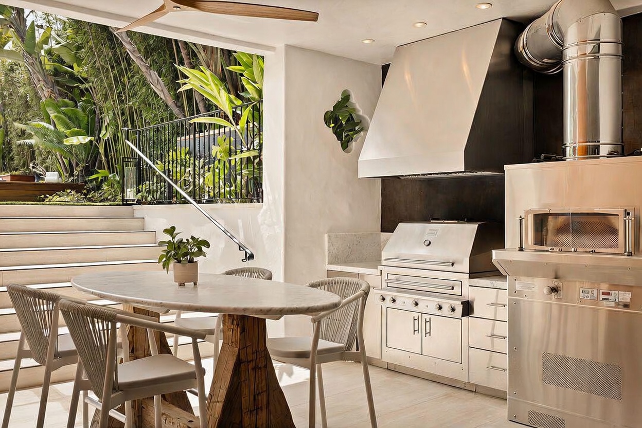 This sophisticated outdoor kitchen and dining area features a stainless steel grill, a built-in pizza oven, and a sleek range hood. An oval stone-topped table with woven chairs sits in the foreground, while a set of stairs leads up to a lush, landscaped garden area. The space is covered, providing a seamless transition between indoor comfort and outdoor living.