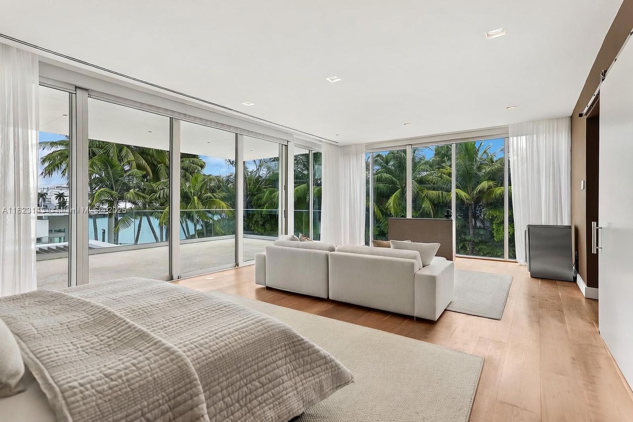 This spacious primary bedroom features light wood flooring and a neutral color palette, centered around a large, plush sofa facing the room. Expansive floor-to-ceiling windows wrap around the space, providing abundant natural light and panoramic views of lush palm trees and the outdoor balcony. The room exudes a serene, modern luxury aesthetic with clean lines and a seamless transition between indoor and outdoor living.