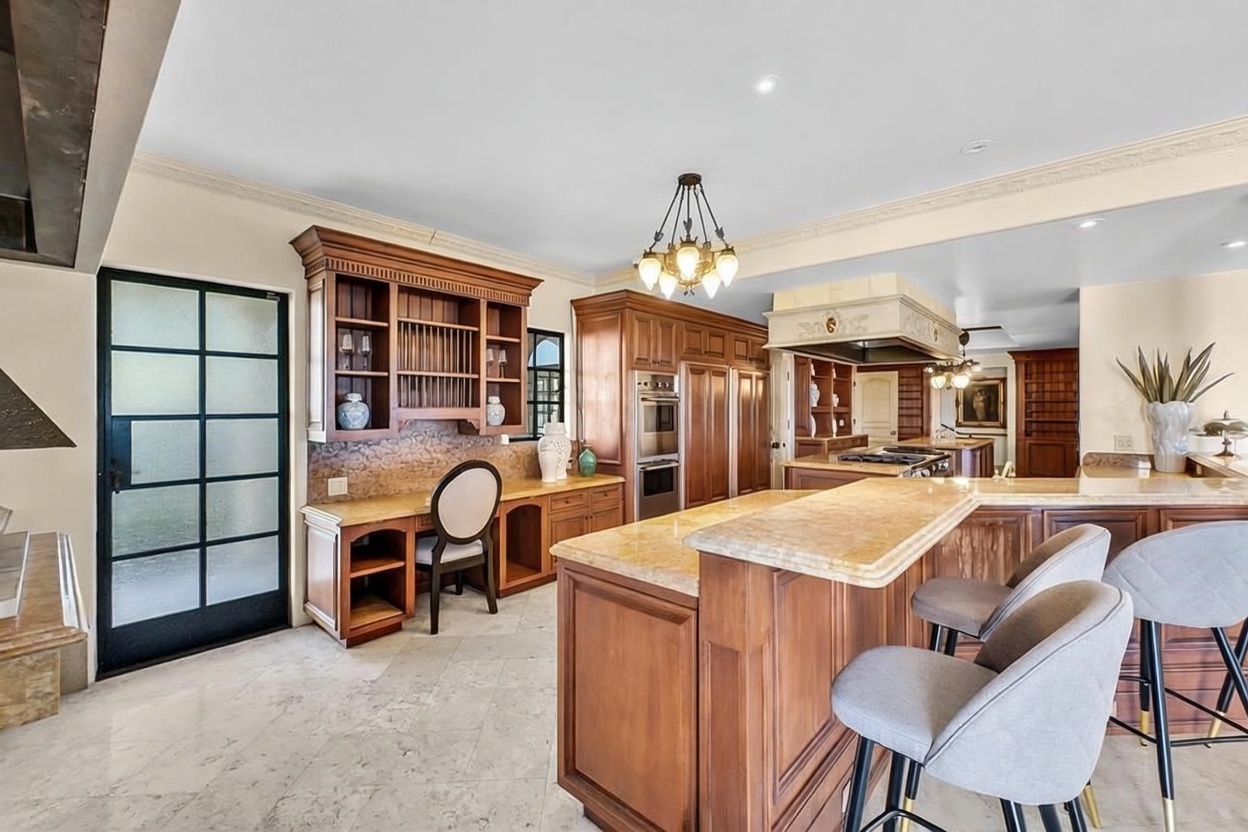 This spacious kitchen features warm wood cabinetry, a large central island with a stone countertop, and a dedicated built-in desk area. The room is illuminated by a decorative chandelier and recessed lighting, with light-colored stone flooring throughout. A black-framed glass door provides a modern contrast to the traditional cabinetry, while bar stools at the island offer casual seating.