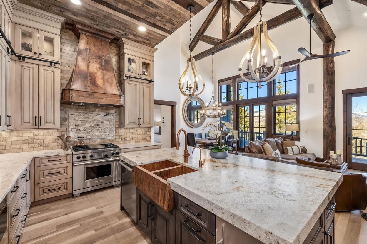 This expansive, rustic-inspired kitchen features a large central island with a copper farmhouse sink and stone countertops, complemented by dark cabinetry and light wood flooring. A prominent copper range hood serves as a focal point above a professional-grade stainless steel stove, while exposed timber beams on the vaulted ceiling add architectural character. The open-concept layout flows seamlessly into a dining and living area, illuminated by large windows that offer views of the surrounding landscape.