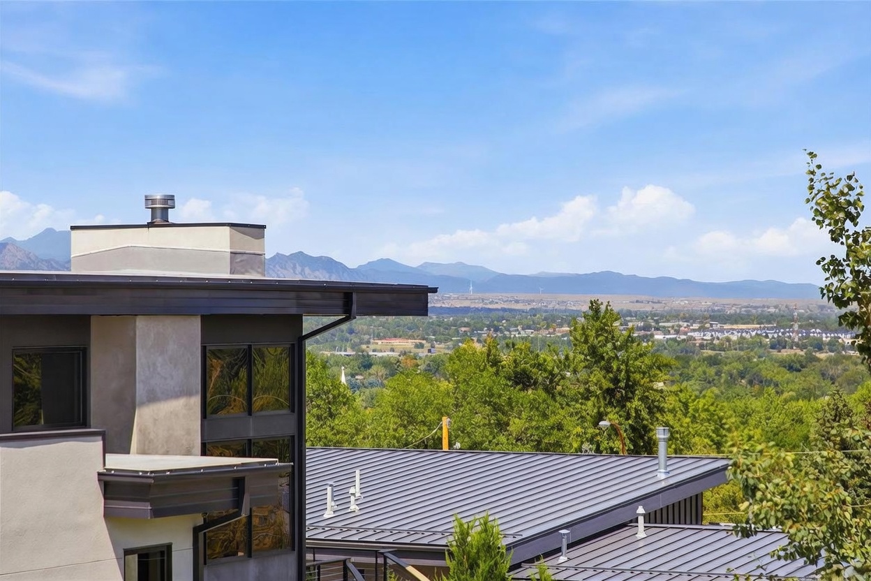 This exterior view captures the modern architectural design of a multi-level home, featuring clean lines, dark metal roofing, and a neutral-toned facade. The perspective highlights the home's elevated position, showcasing expansive, panoramic views of the distant mountain range and the lush valley below. The composition emphasizes the integration of contemporary residential design with the surrounding natural landscape.