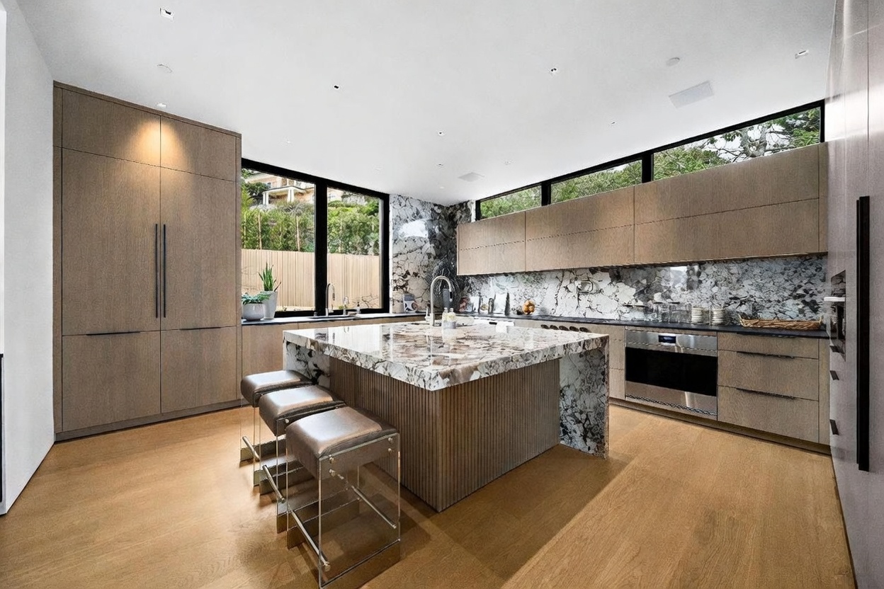 This modern kitchen features a large central island with a stone waterfall countertop and integrated seating for three. The space is defined by light wood cabinetry, a striking patterned stone backsplash, and stainless steel appliances, including a built-in oven. Large windows above the counter and to the side provide ample natural light and views of the outdoors, creating a bright and sophisticated atmosphere.