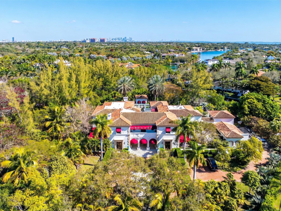 This high-angle aerial view captures a sprawling Mediterranean-style estate nestled within a lush, tropical landscape. The residence features a classic terracotta tile roof, white stucco walls, and a symmetrical facade with arched entryways and red awnings. The property is surrounded by dense greenery and palm trees, with a distant view of a coastal skyline and a nearby waterway.