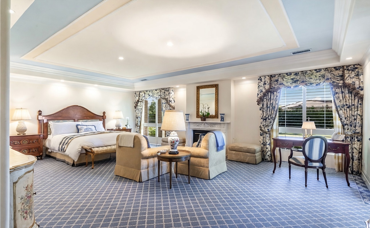 This spacious primary bedroom features a large bed with a wooden headboard, a cozy seating area with two armchairs, and a dedicated desk space by the window. The room is decorated with a classic, traditional aesthetic, highlighted by a tray ceiling, patterned carpeting, and elegant window treatments. A fireplace with a mantel serves as a focal point, adding warmth and character to the expansive suite.