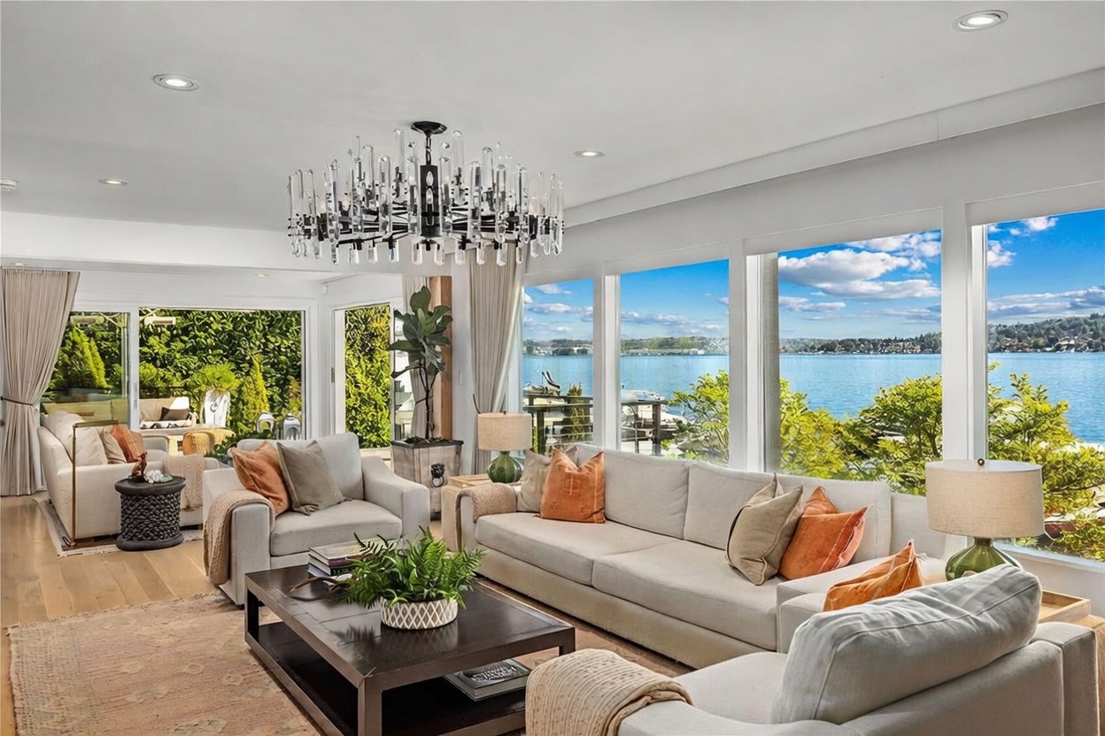 This bright and airy living room features a neutral color palette with light-toned sofas and armchairs accented by warm orange throw pillows. A large, dark wood coffee table sits at the center, while a striking, multi-tiered crystal chandelier serves as a focal point above. The room is defined by expansive windows that provide an unobstructed, panoramic view of the lake and surrounding greenery, creating a seamless connection between the indoor living space and the outdoors.