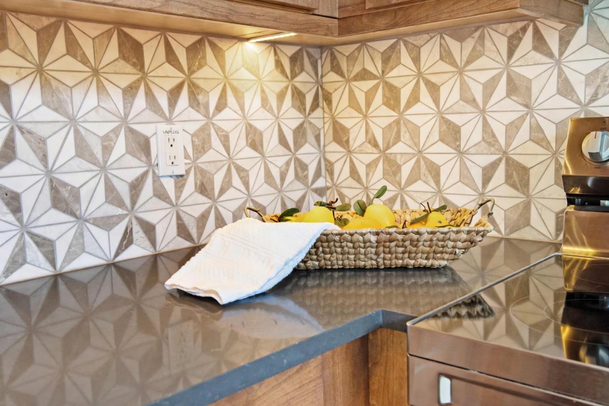 This close-up shot highlights a modern kitchen corner featuring a dark, solid-surface countertop and a striking geometric patterned tile backsplash. A woven basket filled with lemons sits on the counter next to a folded white towel, while an under-cabinet light illuminates the workspace. The scene is framed by warm-toned wood cabinetry and a portion of a stainless steel appliance, creating a clean and inviting culinary aesthetic.