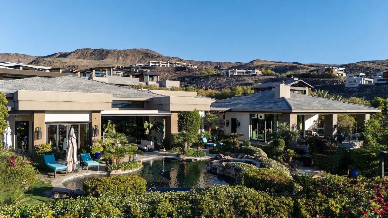 This rear view of a luxury desert residence showcases a sprawling single-story home with a contemporary architectural design, featuring a large swimming pool and lush, manicured landscaping. The property is set against a dramatic mountain backdrop, with expansive glass doors that seamlessly connect the indoor living spaces to the outdoor patio and pool area. The overall impression is one of high-end, tranquil desert living with a focus on indoor-outdoor integration.