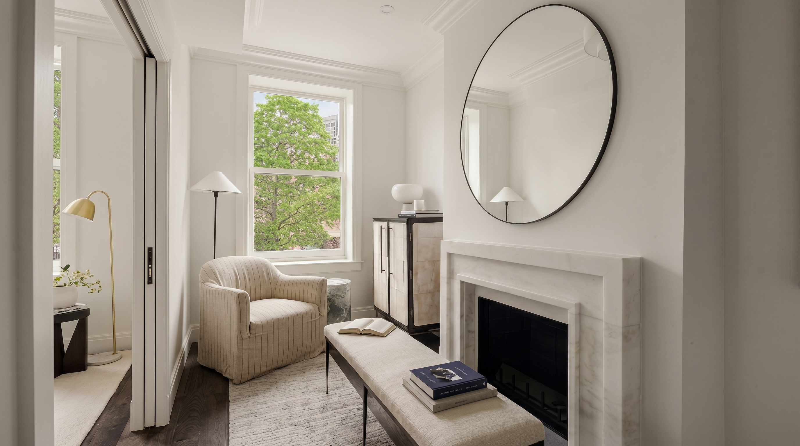 This elegant sitting area features a sophisticated fireplace with a marble surround and a large, circular mirror above it. A plush, neutral-toned bench sits in the foreground on a textured rug, while a cozy striped armchair is positioned near a window that offers a view of lush greenery. The room is finished with dark hardwood floors and a modern ceiling light, creating a serene and refined atmosphere.