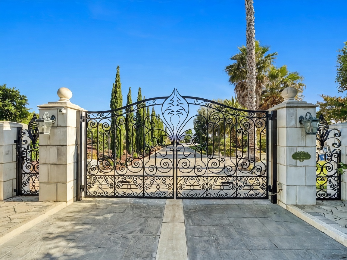 This image captures a grand, ornate wrought-iron gate serving as the primary entrance to a property. The gate is flanked by substantial stone-clad pillars topped with decorative spheres and wall-mounted lanterns, creating a sense of privacy and prestige. The perspective is a straight-on, eye-level shot that emphasizes the symmetry and architectural detail of the entrance, with a paved driveway leading through the open gates toward a sunny, landscaped background.