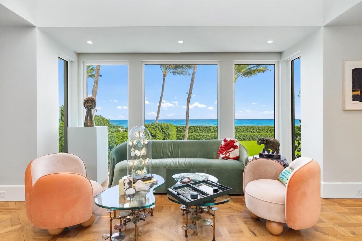 This bright and airy living room features a soft green sofa flanked by two peach-colored armchairs, centered around a pair of glass-topped coffee tables. Large windows provide an expansive, unobstructed view of the ocean, palm trees, and a manicured lawn. The room is accented with unique sculptural decor and warm-toned parquet wood flooring, creating a sophisticated and serene coastal atmosphere.