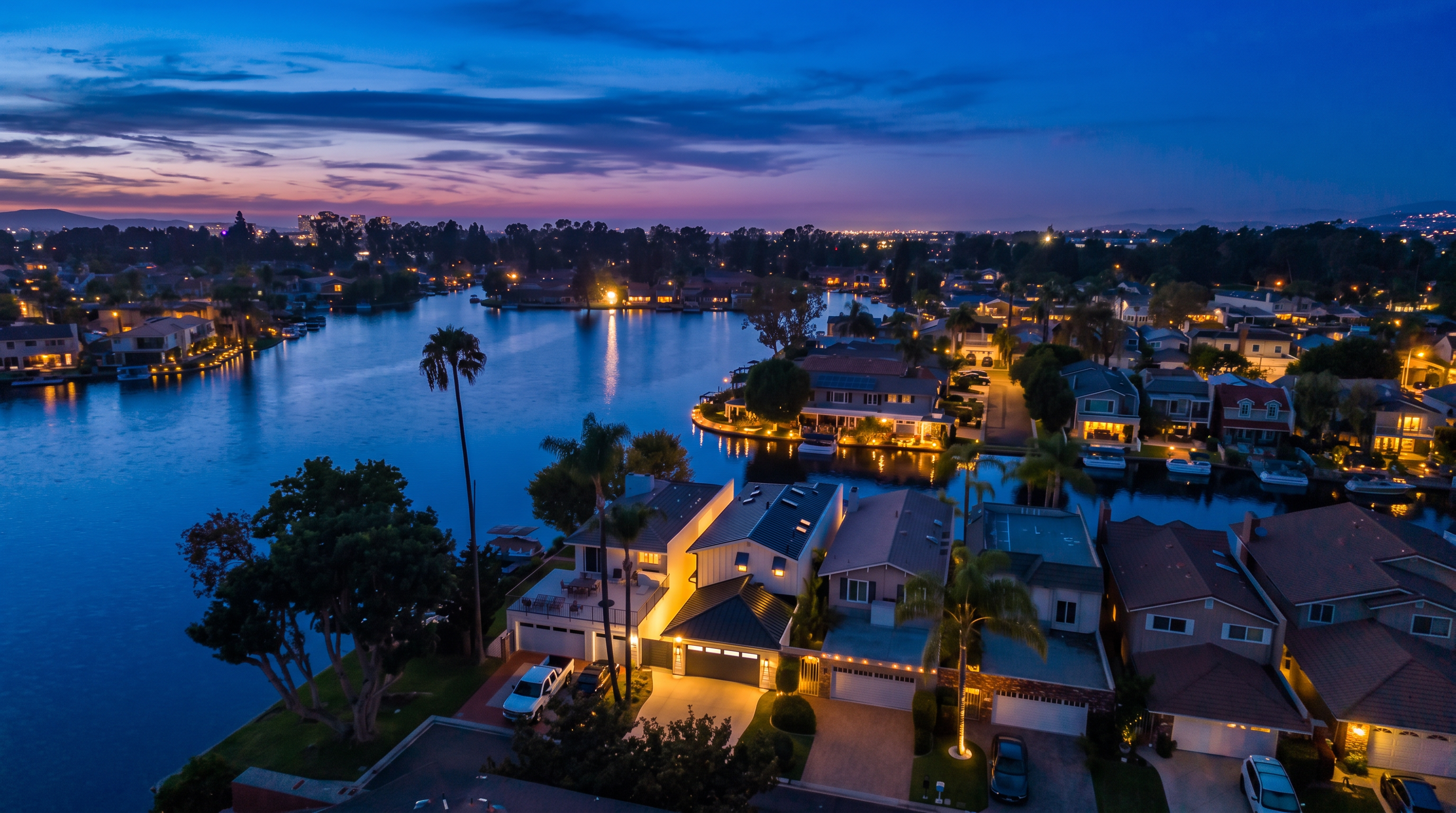 This stunning aerial view captures a waterfront residential neighborhood at sunset, showcasing homes with direct lake access and private docks. The warm golden glow of the sky reflects beautifully off the calm water, highlighting the tranquil and upscale lifestyle of the community. Well-maintained properties with modern architectural styles and manicured driveways create an inviting and prestigious atmosphere.