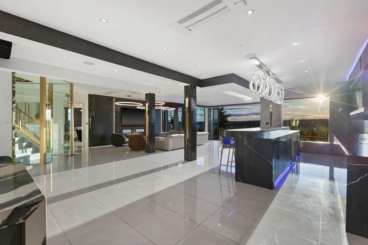This expansive, modern open-concept living space features a sleek kitchen island with a dark stone waterfall edge and integrated blue accent lighting. The room is defined by high-gloss light-colored tile flooring, dark structural columns with gold trim, and a sophisticated lighting scheme including circular pendant fixtures. Large glass doors open to a scenic outdoor view, creating a seamless transition between the luxurious interior and the natural landscape.