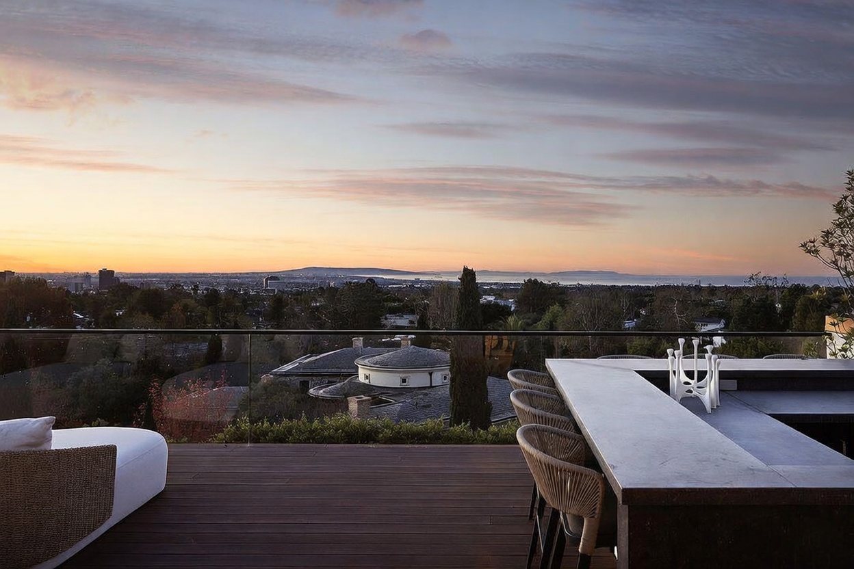 This elevated outdoor deck offers a panoramic sunset view over a suburban landscape and the distant ocean. The space features a sleek, dark-toned bar area with woven chairs and a glass railing that maintains an unobstructed sightline. The warm, golden hour lighting creates an inviting and sophisticated atmosphere for evening relaxation or entertaining.