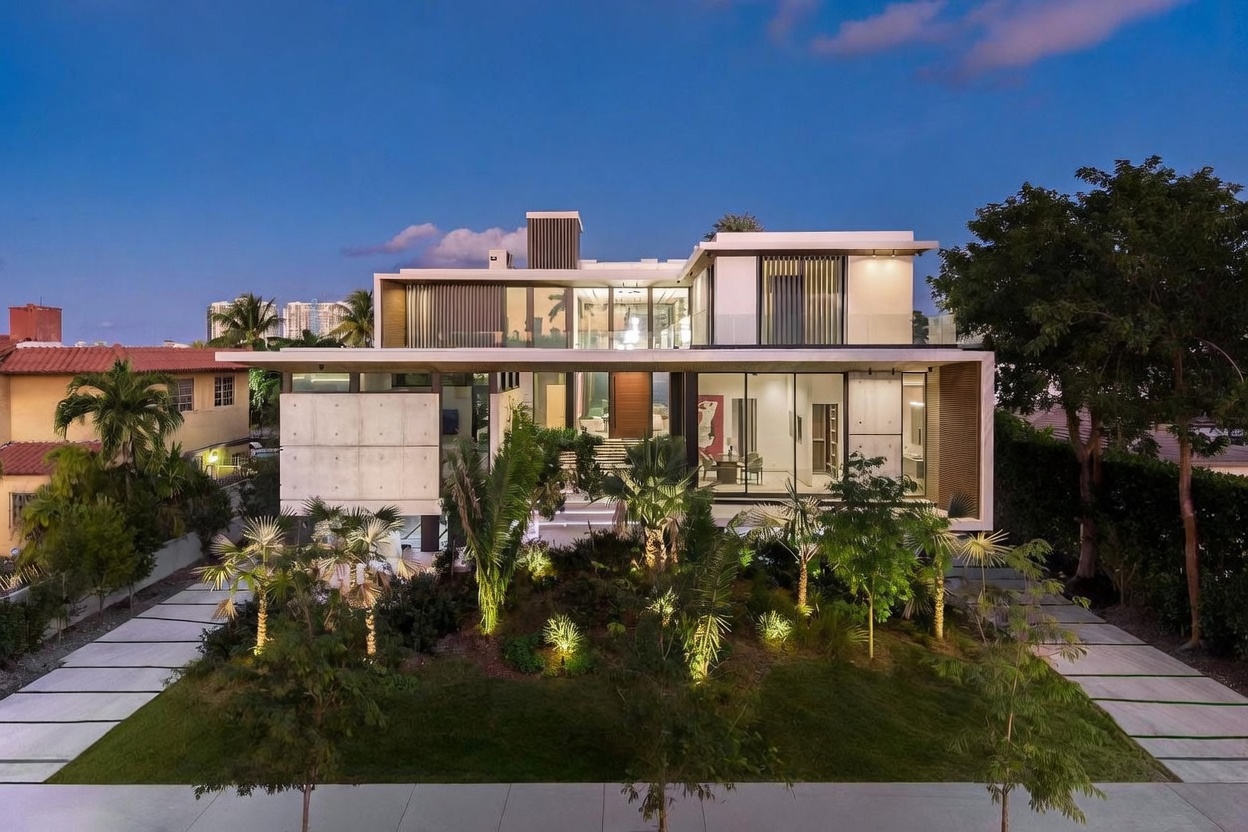 This striking modern residence features a multi-level architectural design with a mix of smooth concrete panels, expansive glass walls, and warm wood accents. The front facade is illuminated by integrated landscape lighting that highlights the lush tropical foliage and palm trees surrounding the property. A wide, paved driveway leads toward the grand entrance, creating a sophisticated and welcoming curb appeal at twilight.