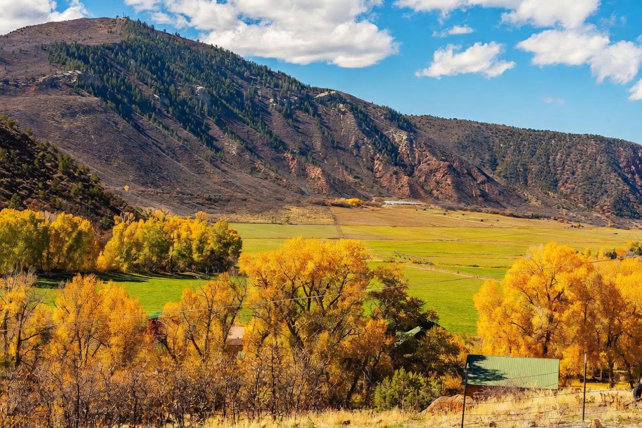 This expansive landscape view captures a vast, golden-hued valley framed by a towering, rugged mountain range under a bright blue sky with scattered clouds. The foreground features vibrant autumn foliage in shades of yellow and orange, surrounding a small structure with a green roof, while the mid-ground showcases open, irrigated green pastures. The perspective is elevated, providing a cinematic, wide-angle look at the property's natural surroundings and rural setting.