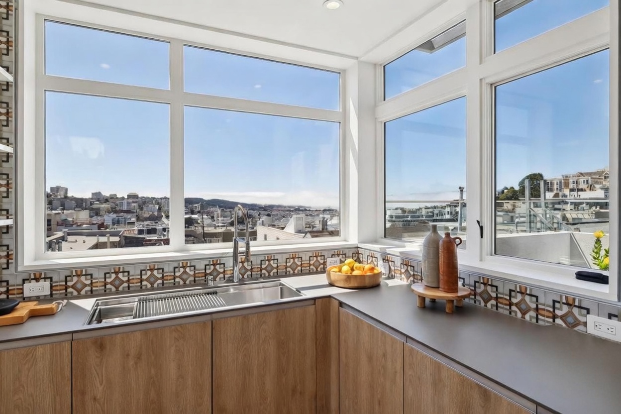 This modern kitchen features a corner sink layout with light wood-toned cabinetry and a dark, matte countertop. A striking backsplash with a geometric, multi-colored pattern runs along the wall, complemented by large windows that provide an expansive, elevated view of the city skyline. The perspective is from a standing position at the sink, highlighting the bright, airy atmosphere and the seamless integration of indoor space with the urban landscape.
