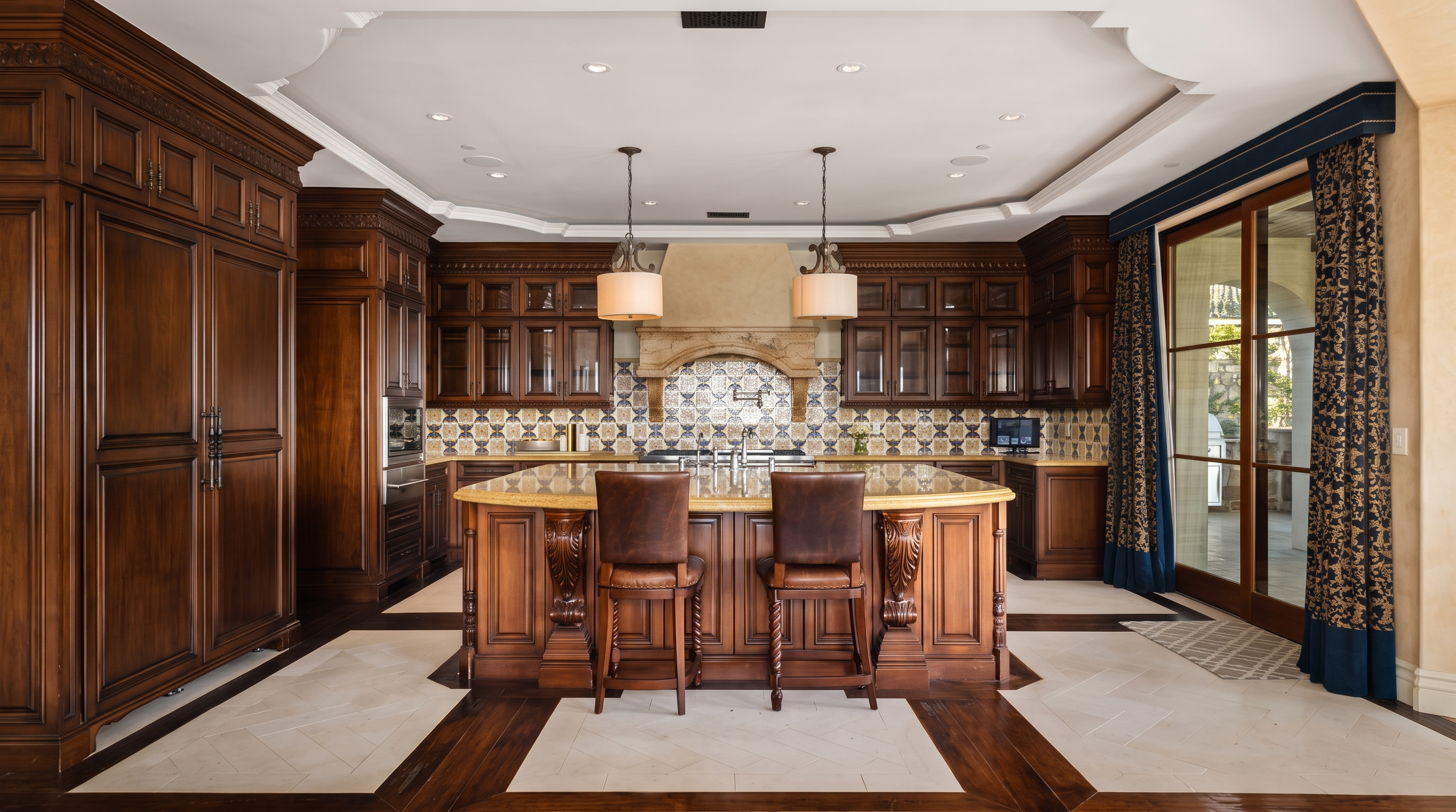 This expansive, traditional-style kitchen features rich, dark wood cabinetry and a large central island with leather-upholstered bar stools. A decorative tile backsplash sits behind the range, complemented by a stone-clad hood, while the floor showcases a sophisticated inlay pattern of light stone and dark wood. The room is framed by a tray ceiling with recessed lighting and large glass doors that lead to an outdoor space, creating a warm and luxurious atmosphere.