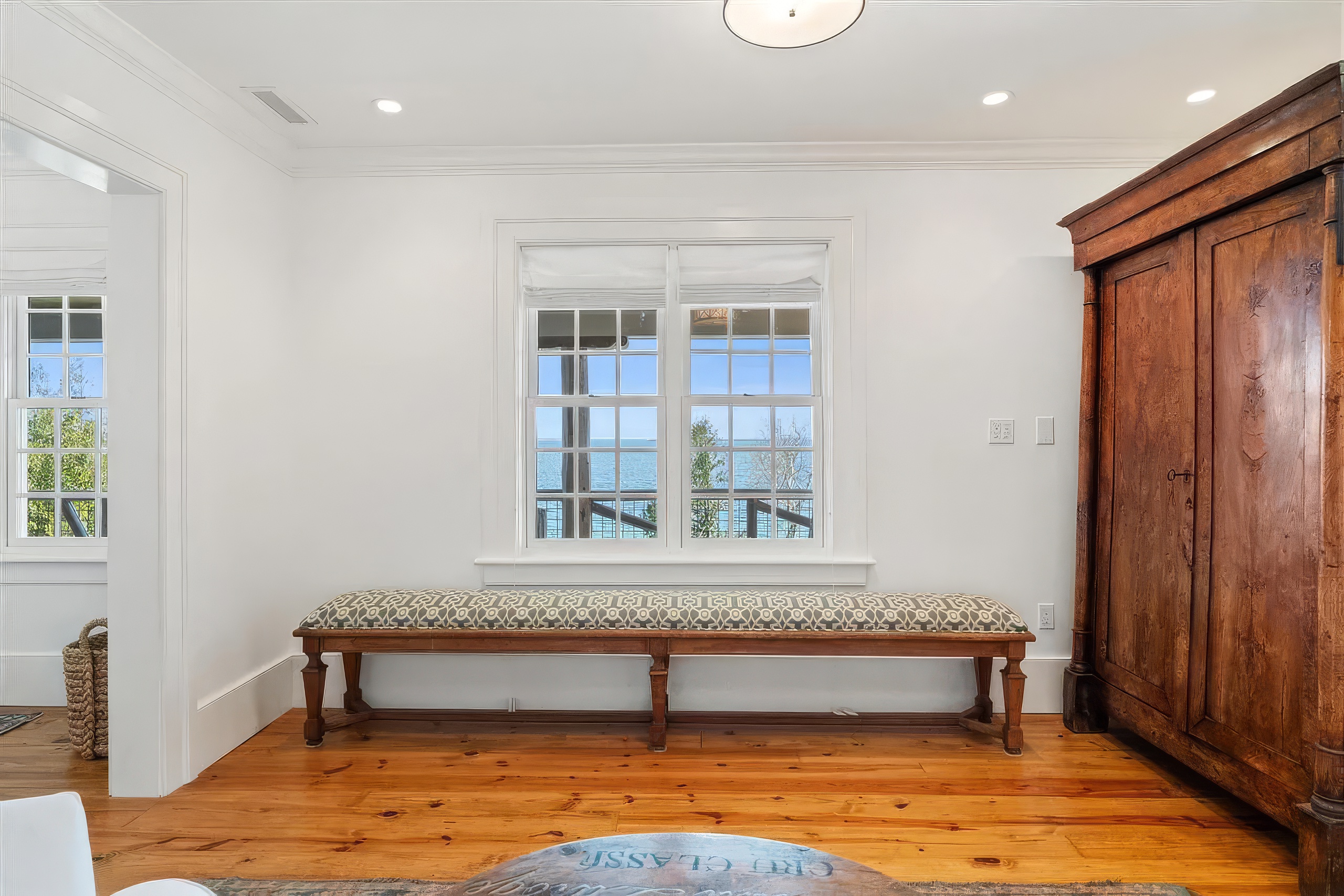 This bright interior space features a long wooden bench with a patterned cushion positioned beneath a large window that offers a scenic view of the water. To the right, a substantial antique-style wooden armoire adds a touch of traditional character, while the room is finished with warm-toned wood flooring and crisp white walls. The perspective is centered on the window, creating a calm and inviting atmosphere suitable for a reading nook or casual seating area.