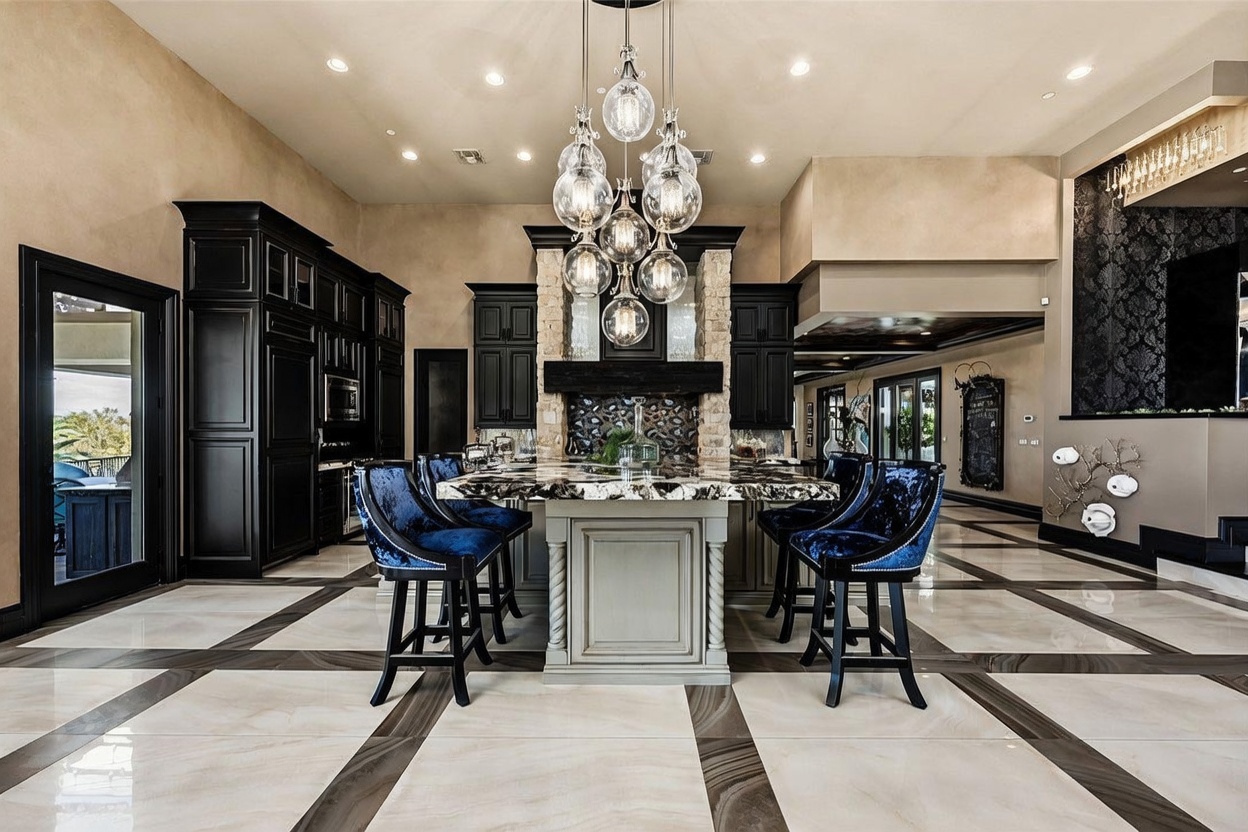 This luxurious kitchen features a large central island with a stone countertop and dark cabinetry, complemented by deep blue velvet bar stools. A striking multi-globe chandelier hangs above the island, while a stone-clad chimney breast serves as a focal point behind the cooking area. The room is finished with polished, large-format patterned tile flooring and recessed lighting, creating a sophisticated and high-end atmosphere.