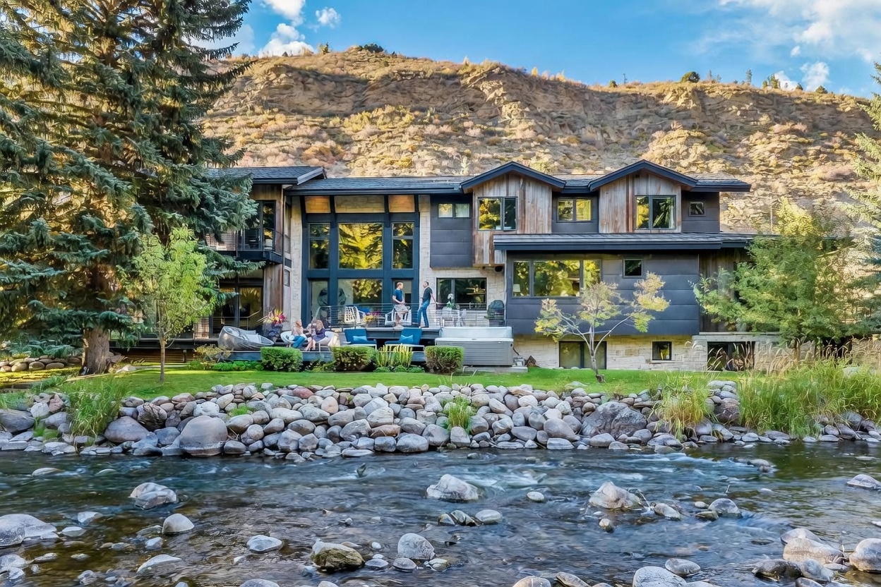 This stunning mountain residence features a contemporary architectural design with a mix of wood siding and dark metal panels, set against a dramatic, rugged hillside. The home includes a spacious multi-level deck overlooking a rushing river, complete with outdoor seating and a hot tub. Large windows and glass doors seamlessly connect the interior living spaces to the natural landscape, creating an inviting and serene atmosphere.