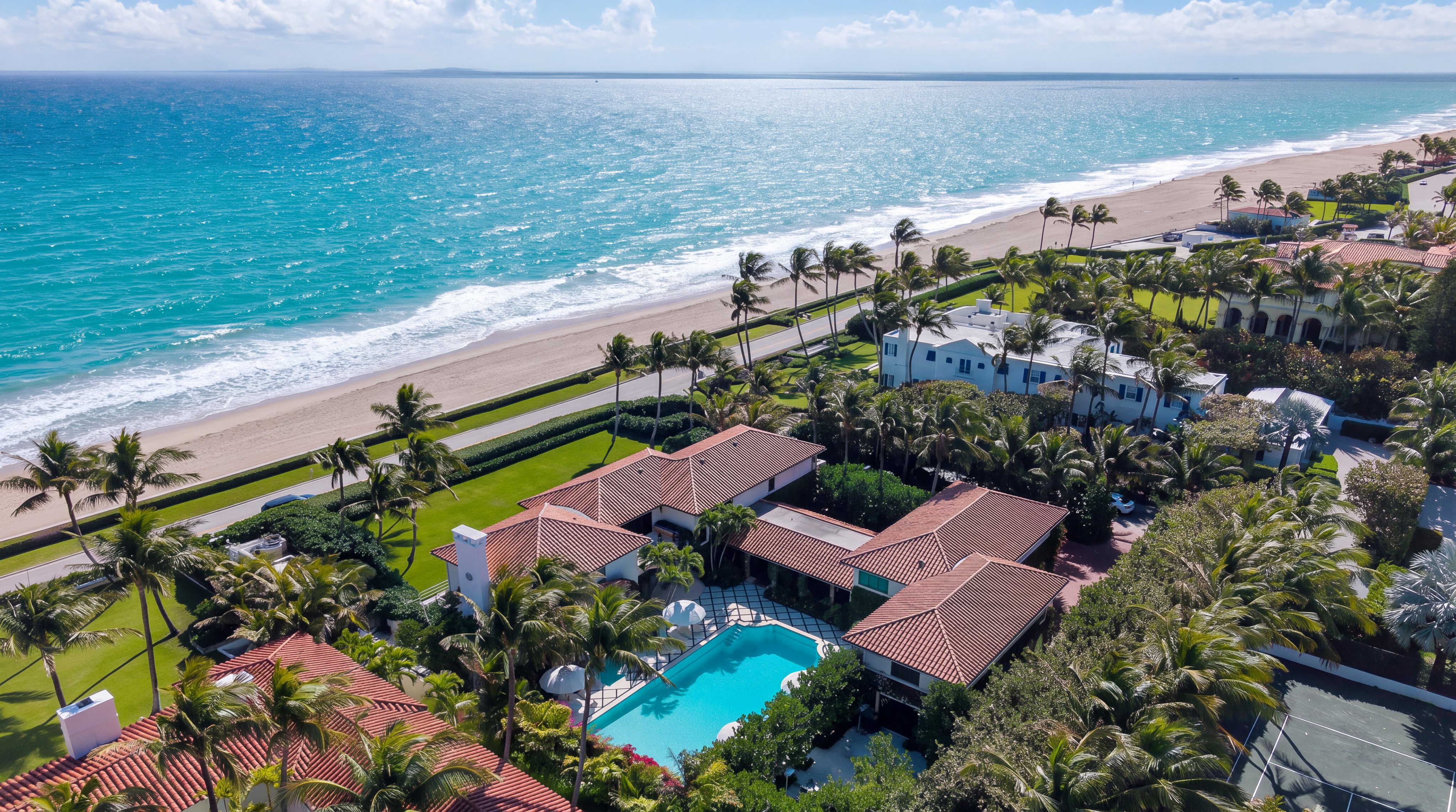This high-angle aerial view captures a sprawling, Mediterranean-style estate situated directly along a pristine coastline. The property features a large swimming pool, a private tennis court, and lush tropical landscaping, all enclosed within a clearly defined boundary. The expansive blue ocean and sandy beach provide a stunning backdrop to this luxurious coastal residence.