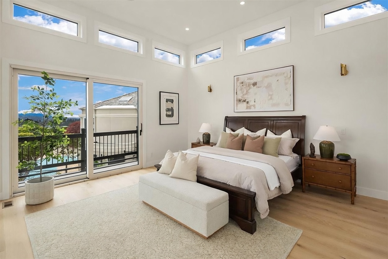 This bright and airy primary bedroom features a dark wood bed frame centered against a white wall, accented by a large piece of abstract art. The room is illuminated by high clerestory windows and a sliding glass door that opens onto a private balcony with a view of the outdoors. A light-colored area rug sits beneath a matching upholstered bench at the foot of the bed, creating a serene and sophisticated atmosphere.