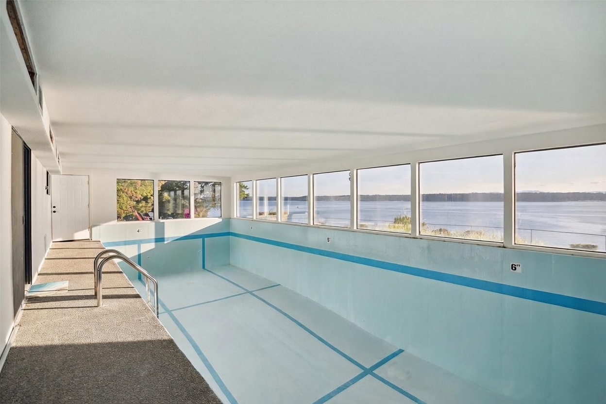 This unique indoor pool room features a series of large windows that provide an expansive, unobstructed view of the water and horizon. The space includes a textured, aggregate-style walkway alongside the pool, which is marked with depth indicators along the wall. The room has a simple, functional design that emphasizes the connection between the indoor swimming area and the scenic outdoor landscape.
