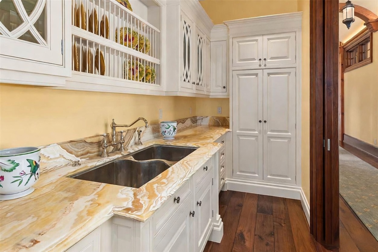 This elegant kitchen area features custom white cabinetry with a built-in plate rack and a tall pantry cabinet. The space is highlighted by a striking, veined stone countertop with an integrated double-basin sink and a polished metal faucet. The warm yellow walls and dark wood flooring create a sophisticated, classic aesthetic, while the perspective looks into the workspace from a doorway leading to a hallway.